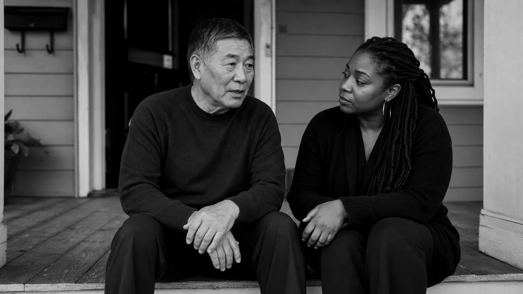 An older East Asian man speaks quietly while a younger Black woman listens beside him on a front porch
