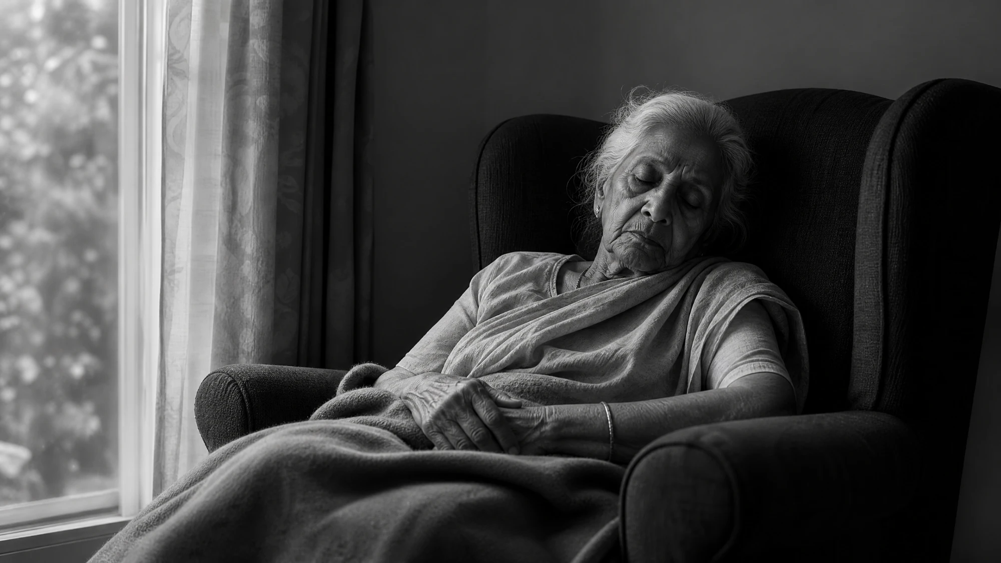 An older South Asian woman resting asleep in a chair by a window, in black and white