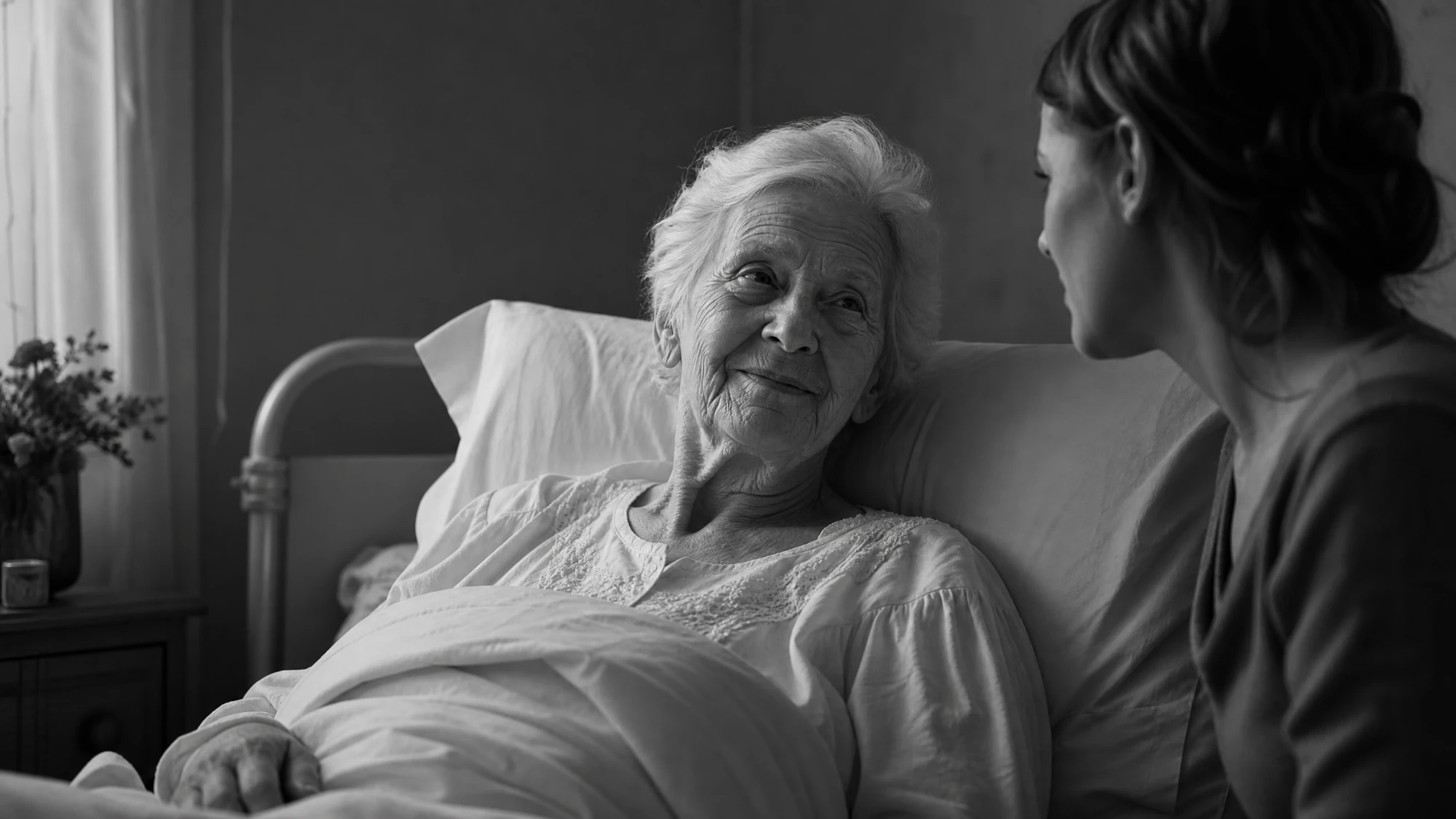 An older woman briefly alert and smiling in bed as a younger woman sits with her, in black and white